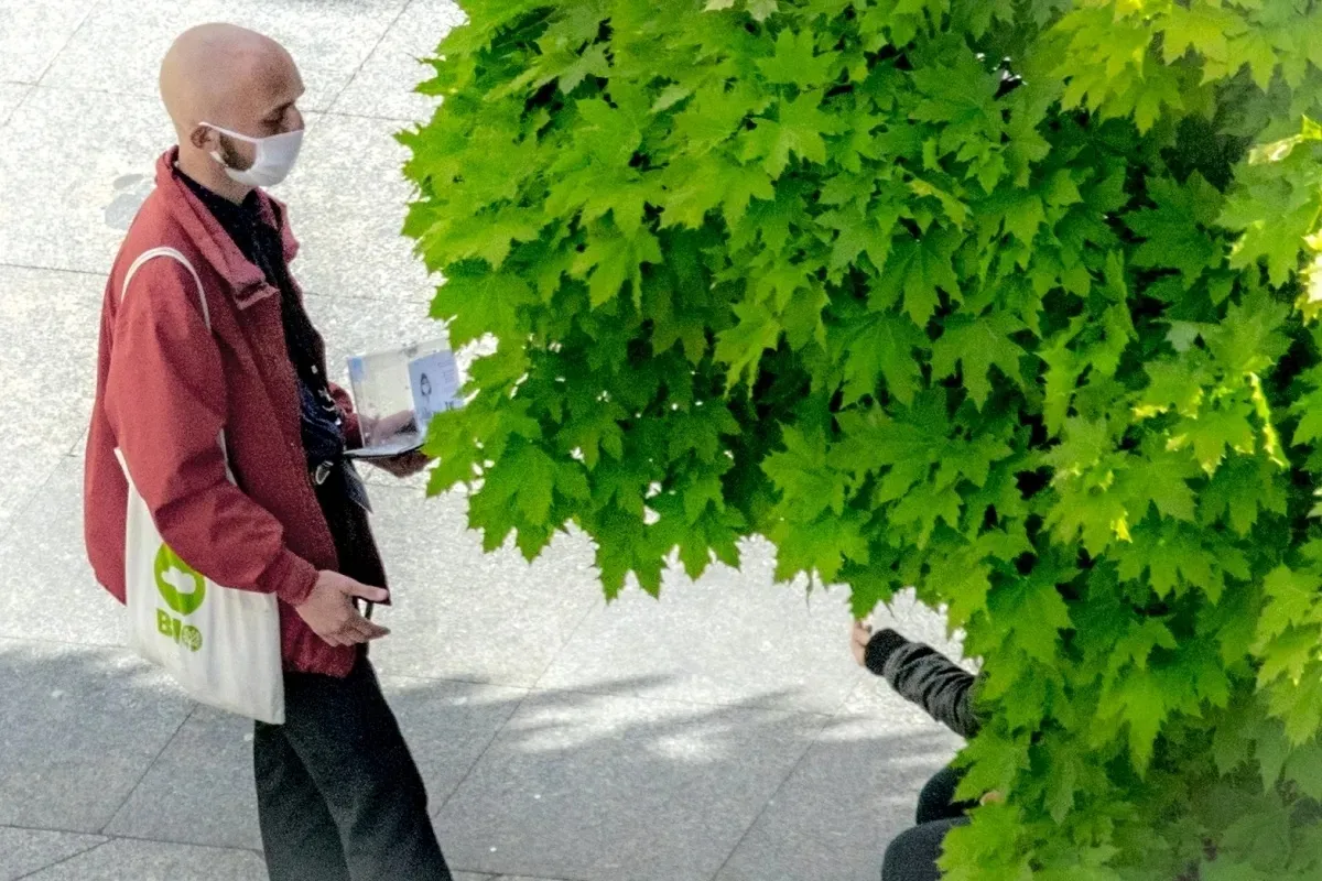 Masked bald man with a tote bag standing beside bright green leaves